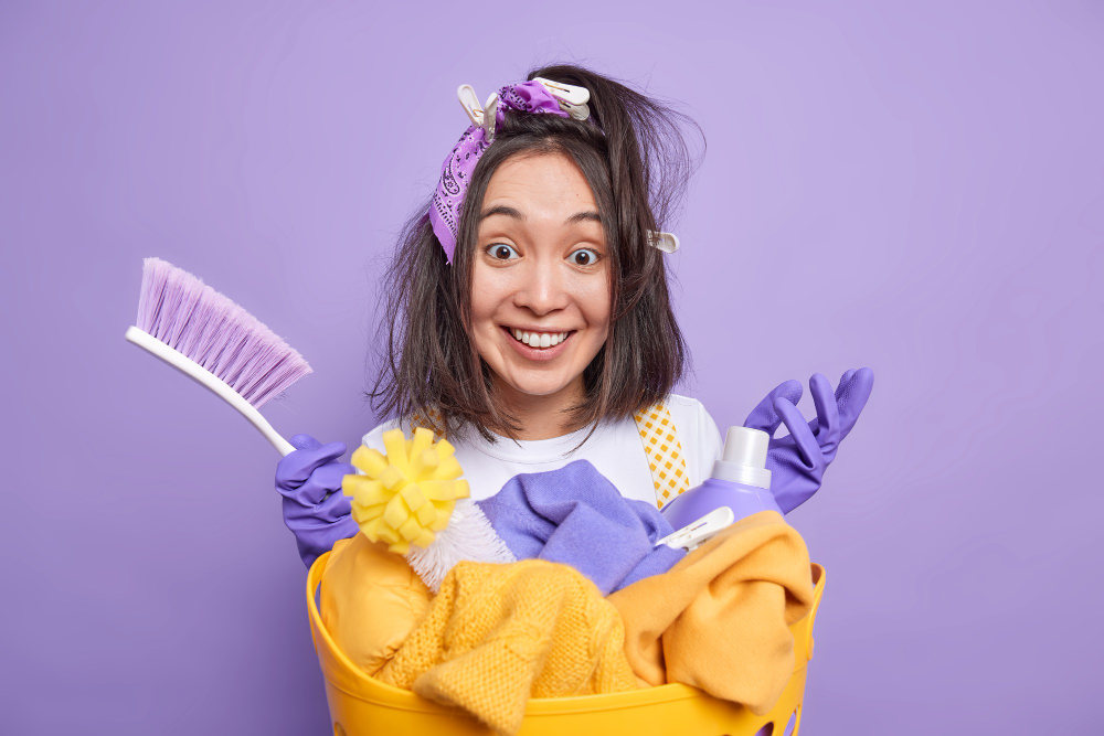 man with clothespins in hair holds brush busy cleaning house smiles positively stands near laundry basket uses detergents isolated on vivid purple